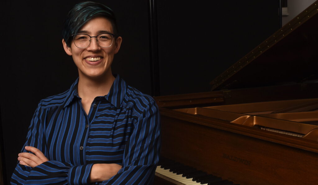 Portrait of Phoebe Wu, piano technician. She is facing the camera with arms folded, in a bright blue striped button down, seated in front of a brown grand piano.