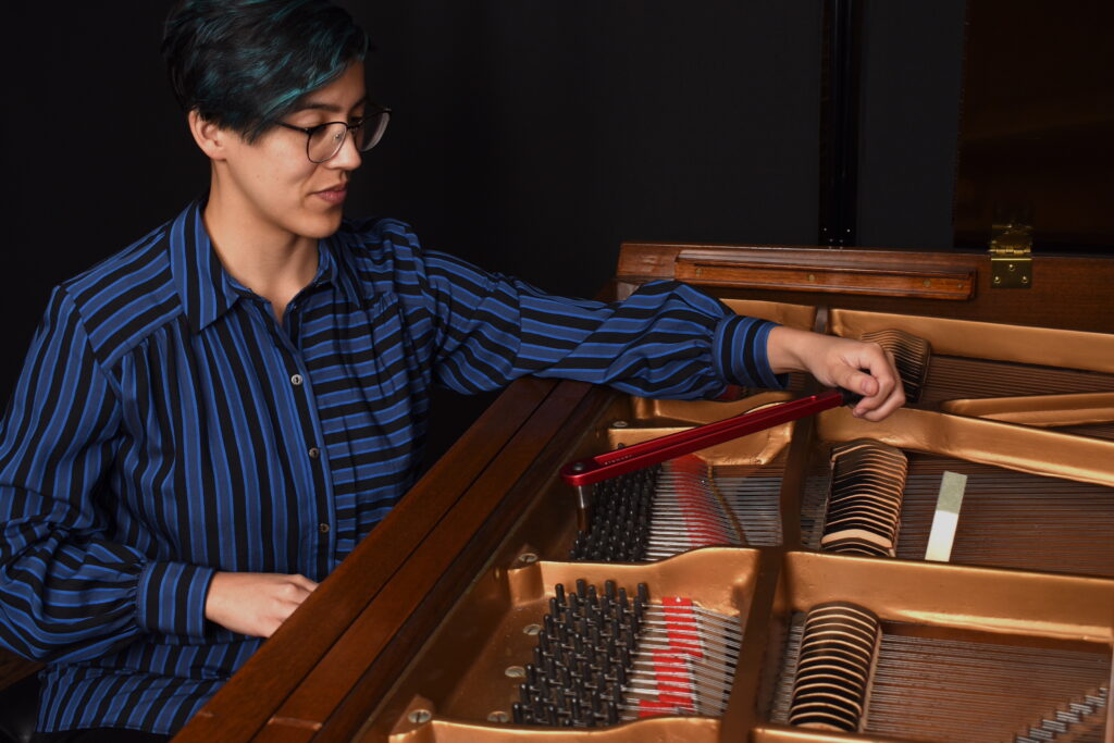 Phoebe Wu, piano technician, is seated at a brown grand piano, looking down at her work. She is wearing a bright blue striped button down and is tuning the piano with a red tuning lever.